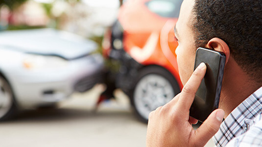 A man holding a cell phone at a scene of a car accident in the background.