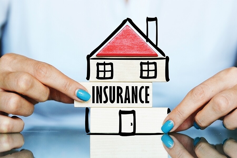 Photo showing two hands with blue fingernail polish holding a small sign in front of a cardboard house which reads: "Insurance".