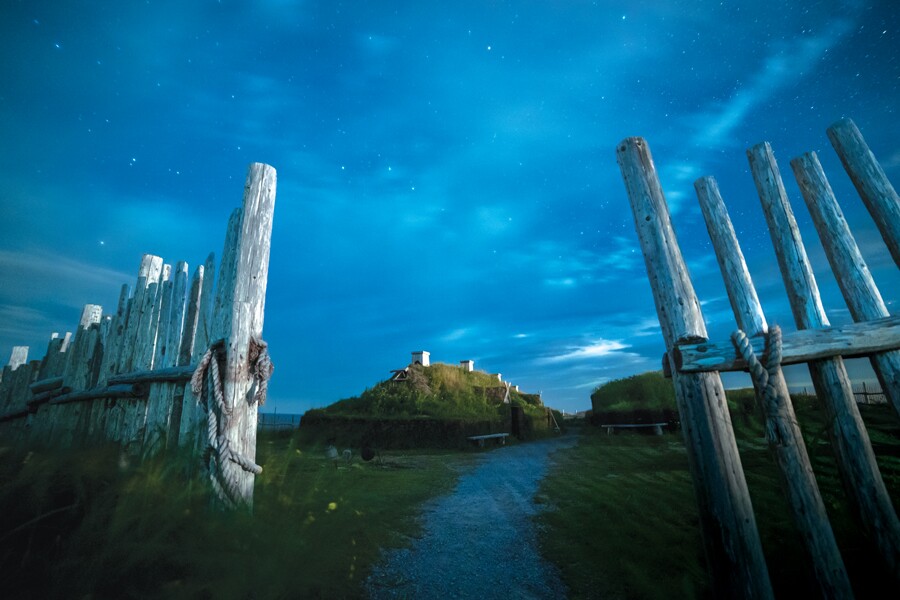 Photo showing old fences in the forground and a parth leading to a small hill with buildings in the far distance.