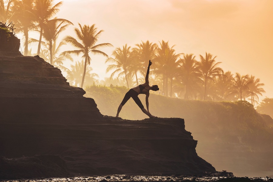 Image showing a person in silhouette doing stretch exercises on a rock outcropping with palm trees in the background.