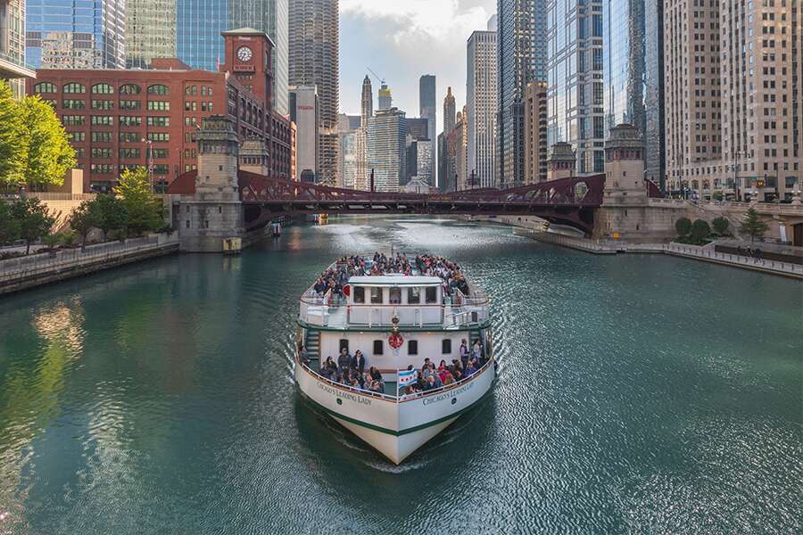 Photo showing a outrist boat sailing past towering office buildings on the Chicago River.