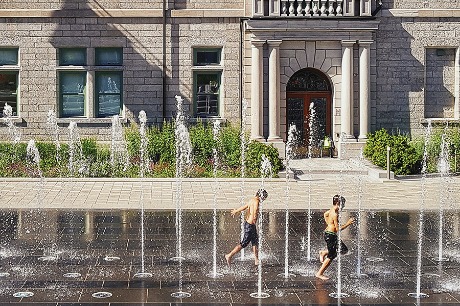 Photo showing two young boys in cut-off jeans running through sprinklers set up outside a large stone building.