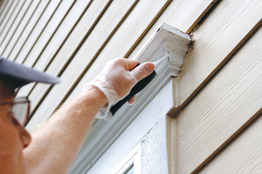 Photo showing partial face of man to the left reaching up and applying caulking to a window frame.