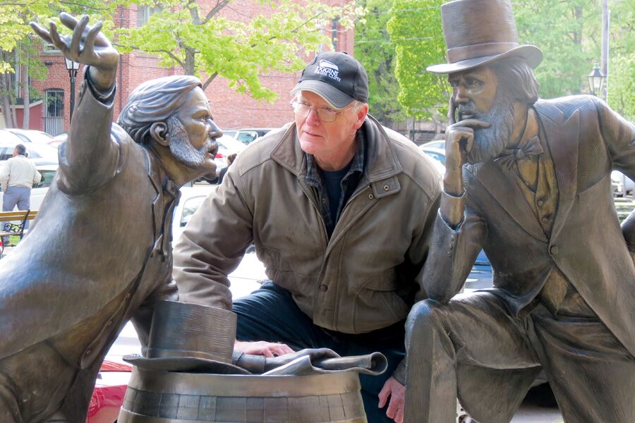 Photo shows a statue of two men engaged in intense debate, with a real man in a blue baseball cap peering at one of the men in the statue.