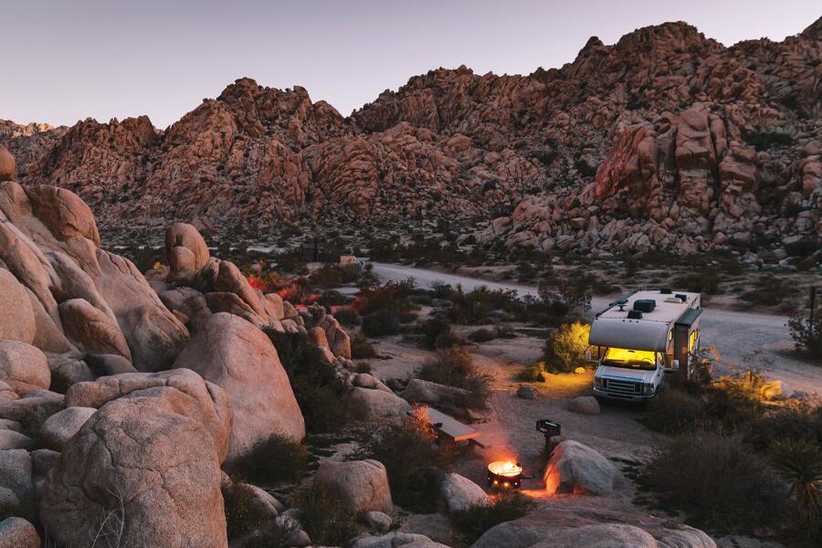 Photo shows an RV parked by campfire off a road running through a small rocky valley.