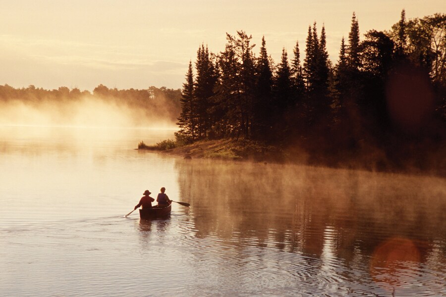 Image shows two people in a canoe paddling near a forested shore with a cloud of mist in the background.