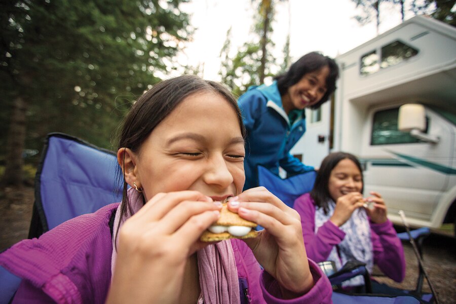 Photo shows two people eating in the foreground with another person looking on and smiling in the background, while sitting beside a parked RV.