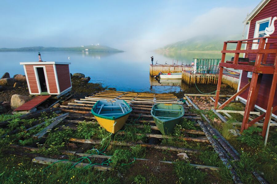 Photo shows two small dory's laying on wood planks beside a red shed building waith a calm harbour in the background.
