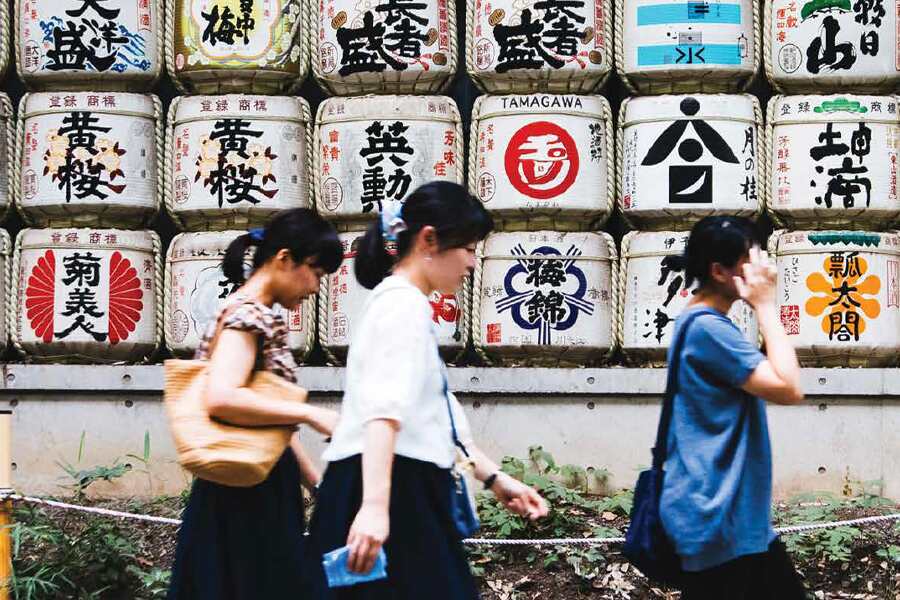Photo showing three young women walking down a street with Japanese writing on a nearby wall.