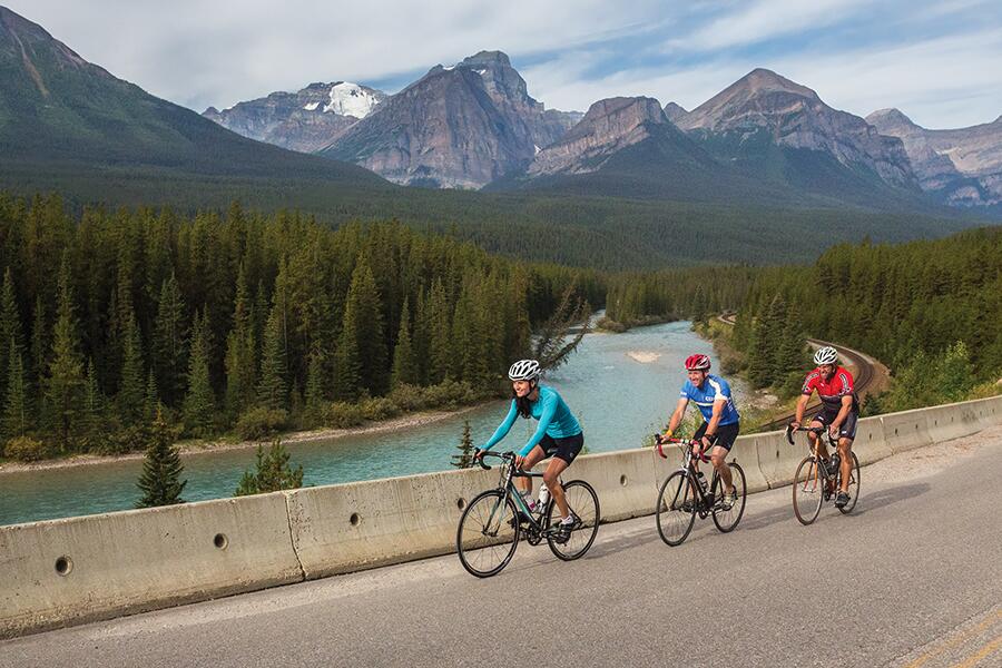 Image showing trio of cyclists along side of highway with river, trees and mountains in background.