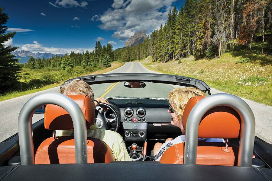 Image showing couple in sports car-like convertible vehicle driving down highway with trees and mountains in background.
