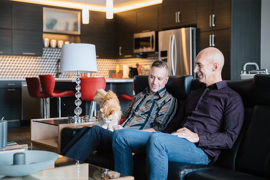 Photo showing two men sitting with cat inside room with stylish, modern furnishings. 