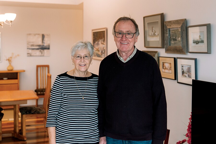 Photo of older couple standing inside home with cozy furniture and family photos on the wall.