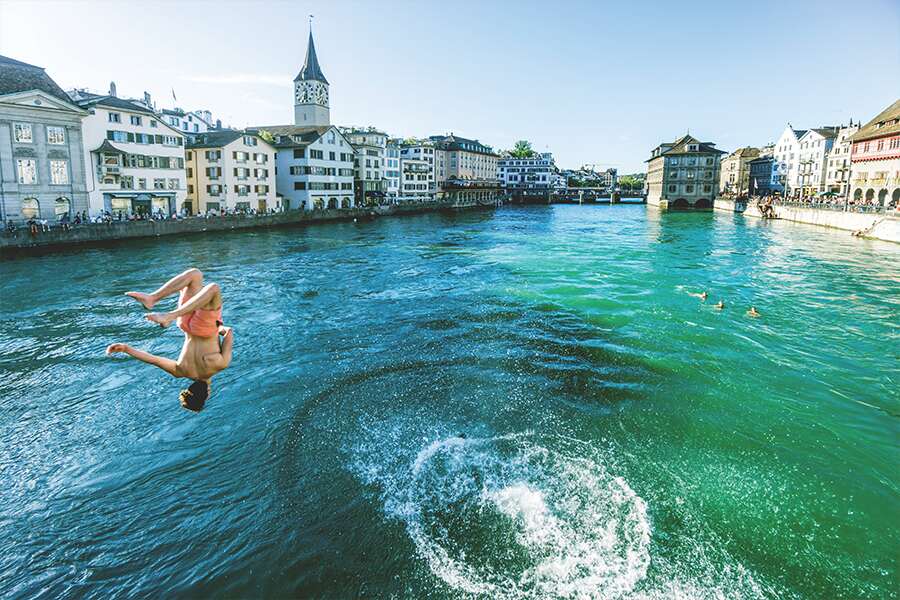 Photo showing young boy somersaulting into picturesque river running through old Zurich.
