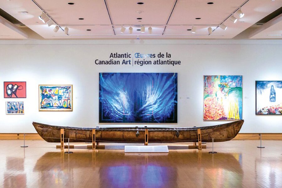 Photo showing hand-made canoe inside the Beaverbrook Art Gallery in New Brunswick with paintings and posters hanging on the wall.