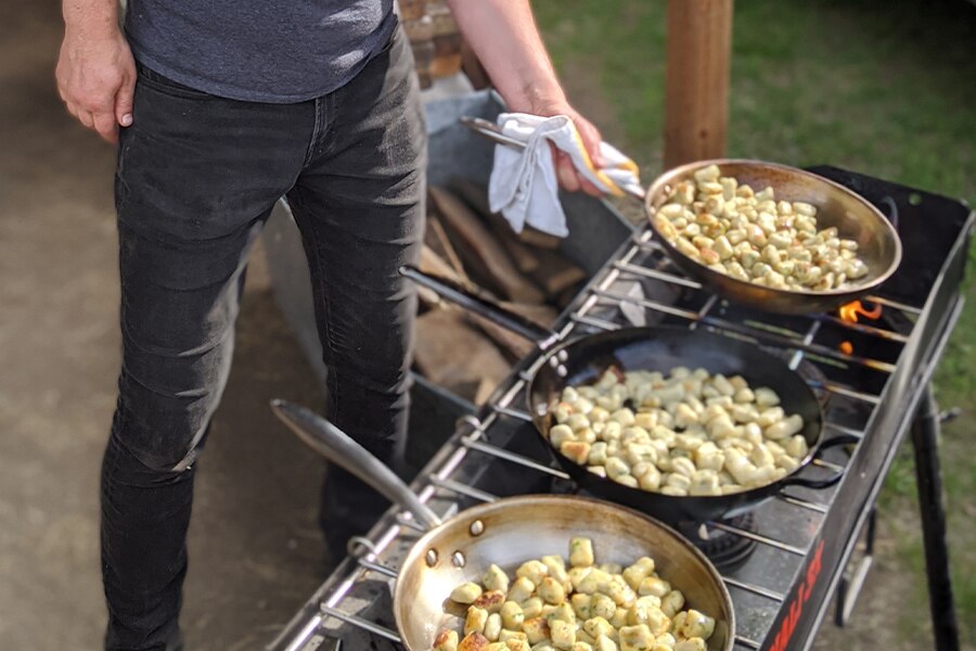 Image shows three skillets of vegetables cooking on an outdoor grill.