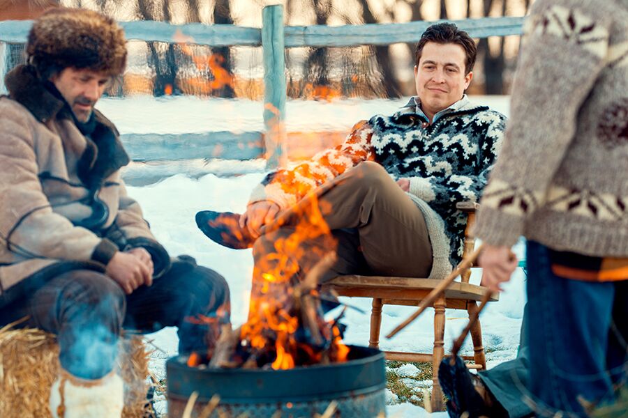 Images showing two men in winter clothing sitting around a camp fire.