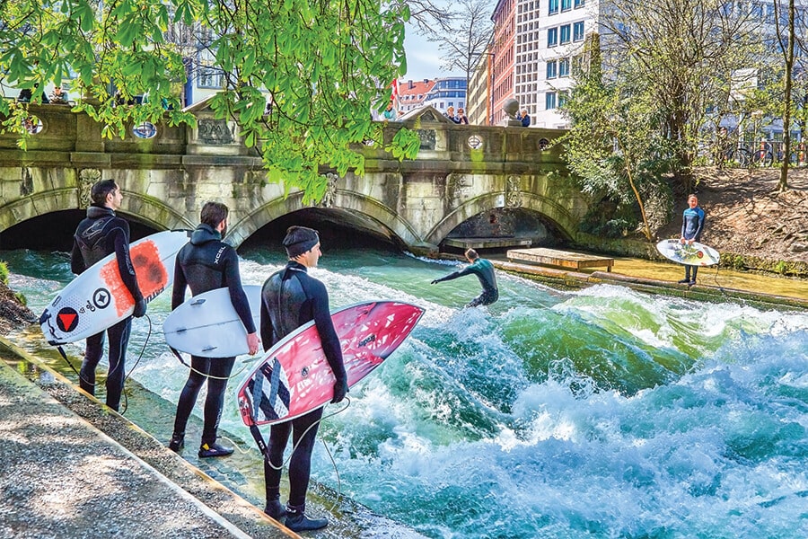 Image showing three men wearing wetsuits and holding surfboards standing on a sidewalk while a fourth man surfs fast-rushing waters in an old canal.