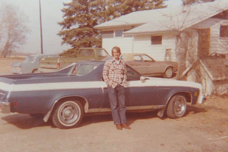 Image showing a man wearing a plaid shirt and blue jeans leaning against a blue and white coloured 1975 Chevrolet El Camino.