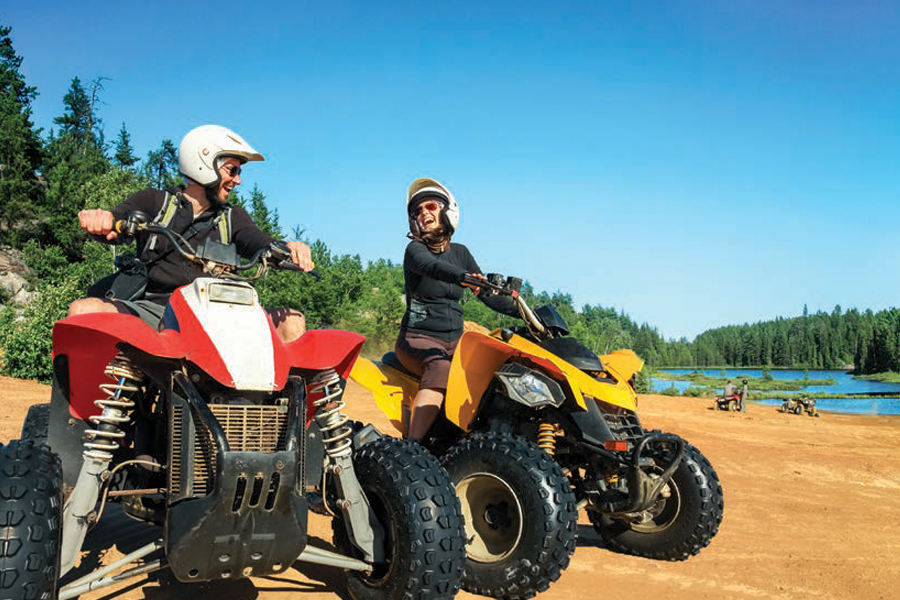 Image showing two people riding all-terrain vehicles beside a lake with a forest in the background.