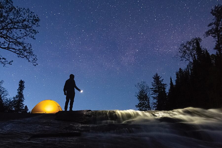 Image showing a night time view of a starry rural sky, with a man standing near a lit up tent holding a flashlight.
