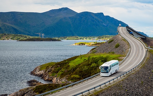 A motorcoach on a road overlooking a lake with a mountain in the background.