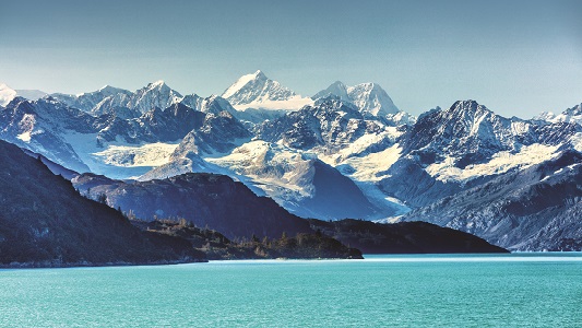 View of snow-covered Alaskan mountains with turquoise green ocean in foreground.