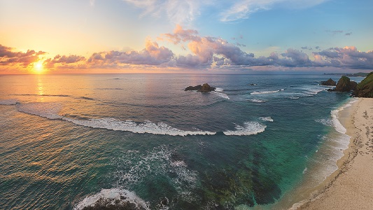 View of ocean and sandy beach with a setting sun in the background.