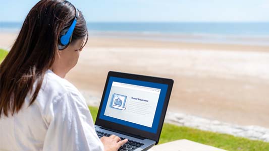 Woman sitting on beach with headphones using laptop to research CAA Travel Insurance