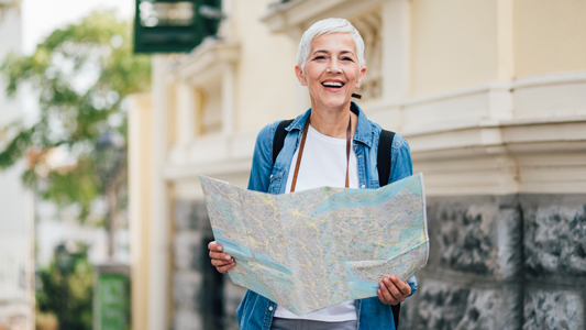 Image showing smiling woman with short white hair wearing a blue jacket standing outside a building with a map unfolded in her hands.