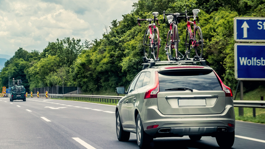 A grey SUV with three bicycles on top driving down a highway alongside trees.