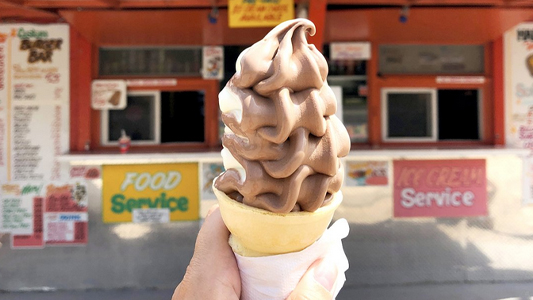 Image showing a hand holding up a chocolate ice cream cone with the ice cream shop in the background.