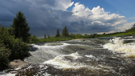 Image showing roiling water on a fast moving river with pine trees to the left and a stormy sky in the background.
