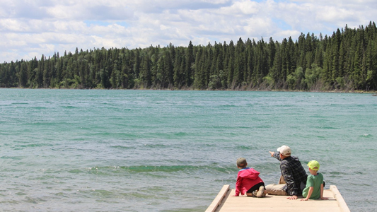 Images showing a man and two children sitting on a dock beside a lake with forest in the background.