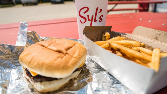 Images showing a hamburger and a box of french fries on a table with a small label reading Syl's.