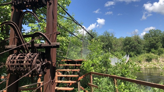 Image showing an old piece of machiney rusting away beside a small lake with a swinging bridge in the background.