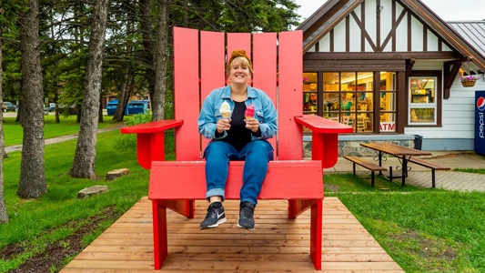 Image showing a woman sitting in a large red wooden chair holding two ice cream cones with a chalet-style building in the background.