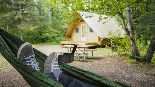 Image showing two feet in a hammock in the foreground with a small wooden building in the background.