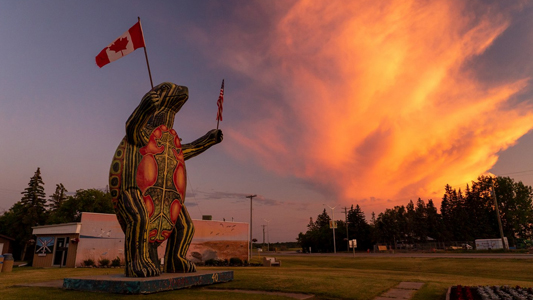 Image of a statue of a large stylized turtle holding up Canadian and American flags under a fiery evening sky.