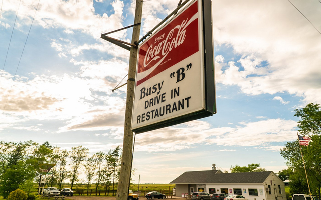 Image showing a sign in the foreground with a Coca Cola logo and words reading Busy B Drive In restaurant and a white building in the background.