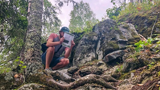 Image showing a man squatting down in the woods beside a large rock and reading a magazine.