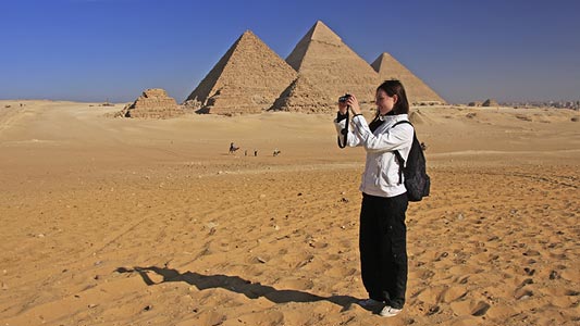 A woman standing in a desert taking a picture in front of the pyramids.