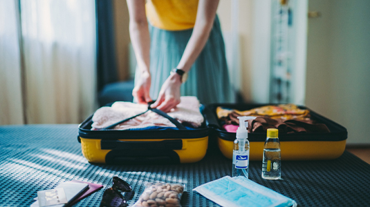 A female traveller packing a suitcase, with face masks, hand sanitizer, and other personal items placed on a bed. 