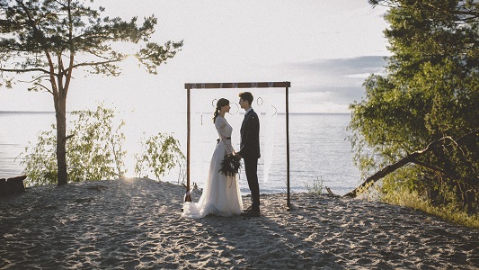 Photo shows wedding couple standing inside a wooden frame beside a lake in summer.
