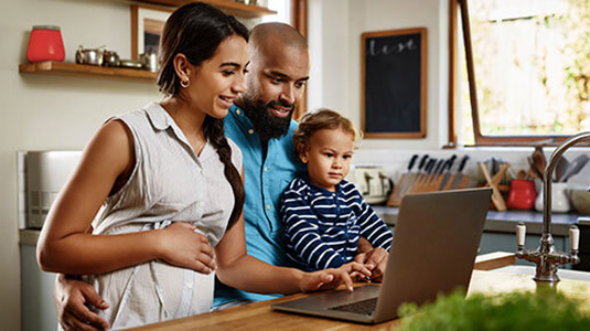 A family of three gathered around a kitchen counter looking at a laptop screen.