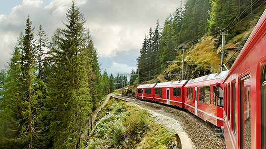 Red train passing on a cliff through a beautiful forest.